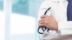A doctor wearing a white coat and holding a stethoscope across their chest, standing in a bright medical office.